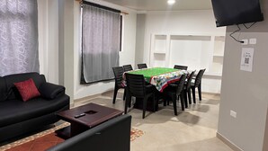 Un salon comprenant un canapé, une table à manger avec des chaises, un téléviseur fixé au mur et une fenêtre avec des rideaux.