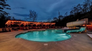 Une piscine avec un toboggan, entourée de chaises longues et d’un espace de détente couvert agrémenté de guirlandes lumineuses.