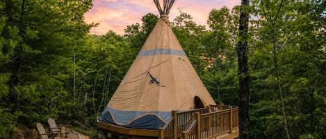 Una tienda de campaña tipi con una terraza de madera y mesas de picnic en un entorno boscoso.