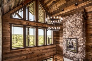L’intérieur d’une cabane en rondins, avec de grandes fenêtres, une cheminée en pierre et un lustre.