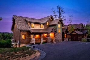 A large, multi-story log cabin with a stone foundation, multiple windows, and a well-lit entrance.