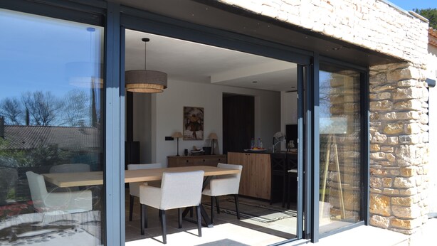 A modern kitchen with a dining area visible through glass doors, a stone exterior wall, and a view of the outdoors. - Small house near Uzes and the Pont du Gard, ideal for 2 or 4 people (Saint-Maximin)