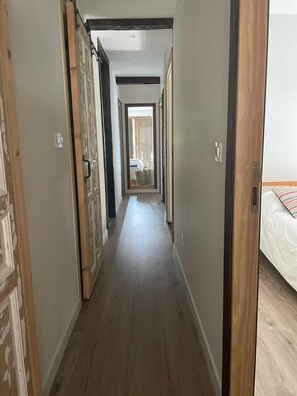 Un couloir étroit, doté d’un plancher en bois, d’où l’on aperçoit une pièce contenant un lit.