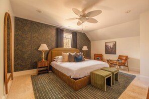 Une chambre avec un grand lit, deux tables de chevet avec des lampes, une chaise, une petite table, un ventilateur de plafond et une fenêtre avec des rideaux.