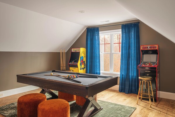 Une table de billard avec des tabourets, un jeu de fléchettes et un flipper dans une pièce dotée d’un plafond en pente et de grandes fenêtres.