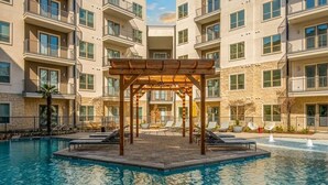 A multi-story building with balconies, a pool area with a wooden pergola, and lounge chairs. - Cozy Designer Suite | Gym & Pool | DART Access (Irving)