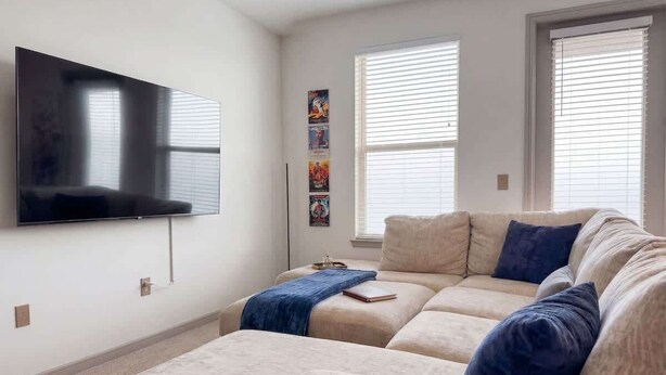 A living room with a large flat-screen TV mounted on the wall, a beige sectional sofa with blue cushions, and a small coffee table. - Cozy Designer Suite | Gym & Pool | DART Access (Irving)