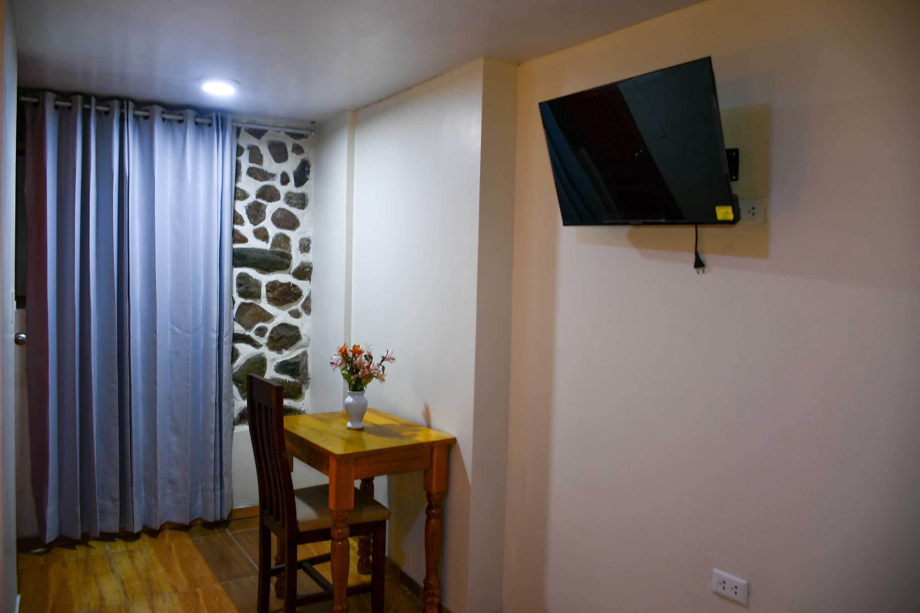 A room with a stone wall, a wooden table with a vase, a TV mounted on the wall, and a window with curtains.