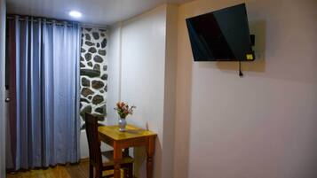 A room with a stone wall, a wooden table with a vase, a TV mounted on the wall, and a window with curtains.