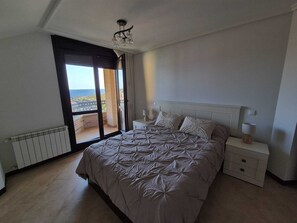 Ett sovrum med en stor säng, ett nattduksbord, en lampa och utsikt över havet genom en skjutdörr.