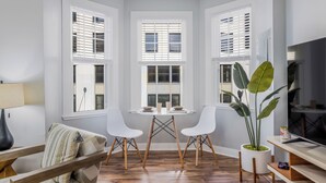 Una sala de estar moderna con una mesa de comedor, dos sillas blancas, un sofá, un televisor y ventanas grandes con persianas.