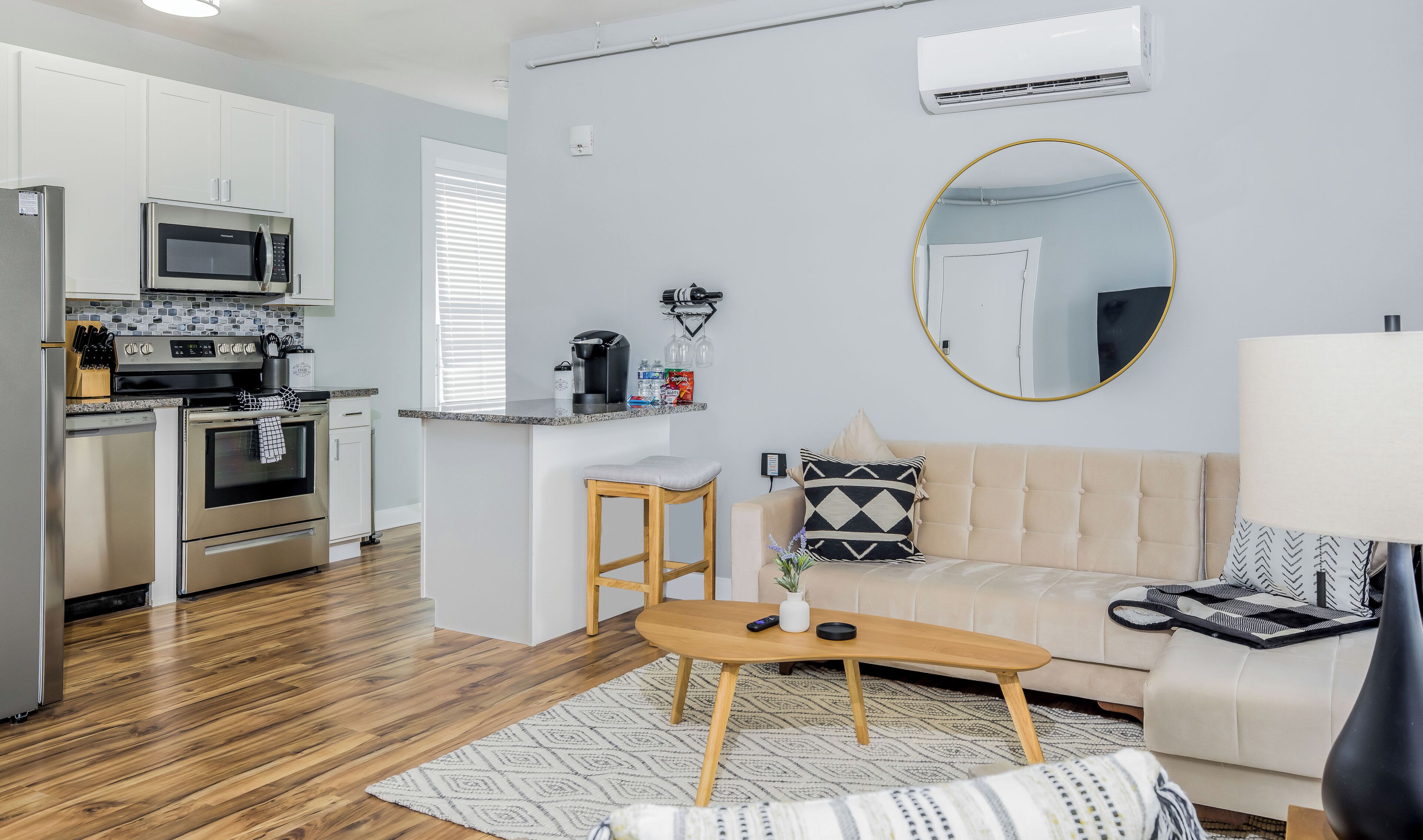 A modern kitchen with stainless steel appliances, a dining area with a wooden table and bar stools, and a living room with a sofa and a round mirror.