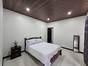 A bedroom with a wooden ceiling, a bed with a white bedspread, a small bedside table with a plant, a door, and a mirror. - Entire New House Hosted by Arelis (San Isidro)