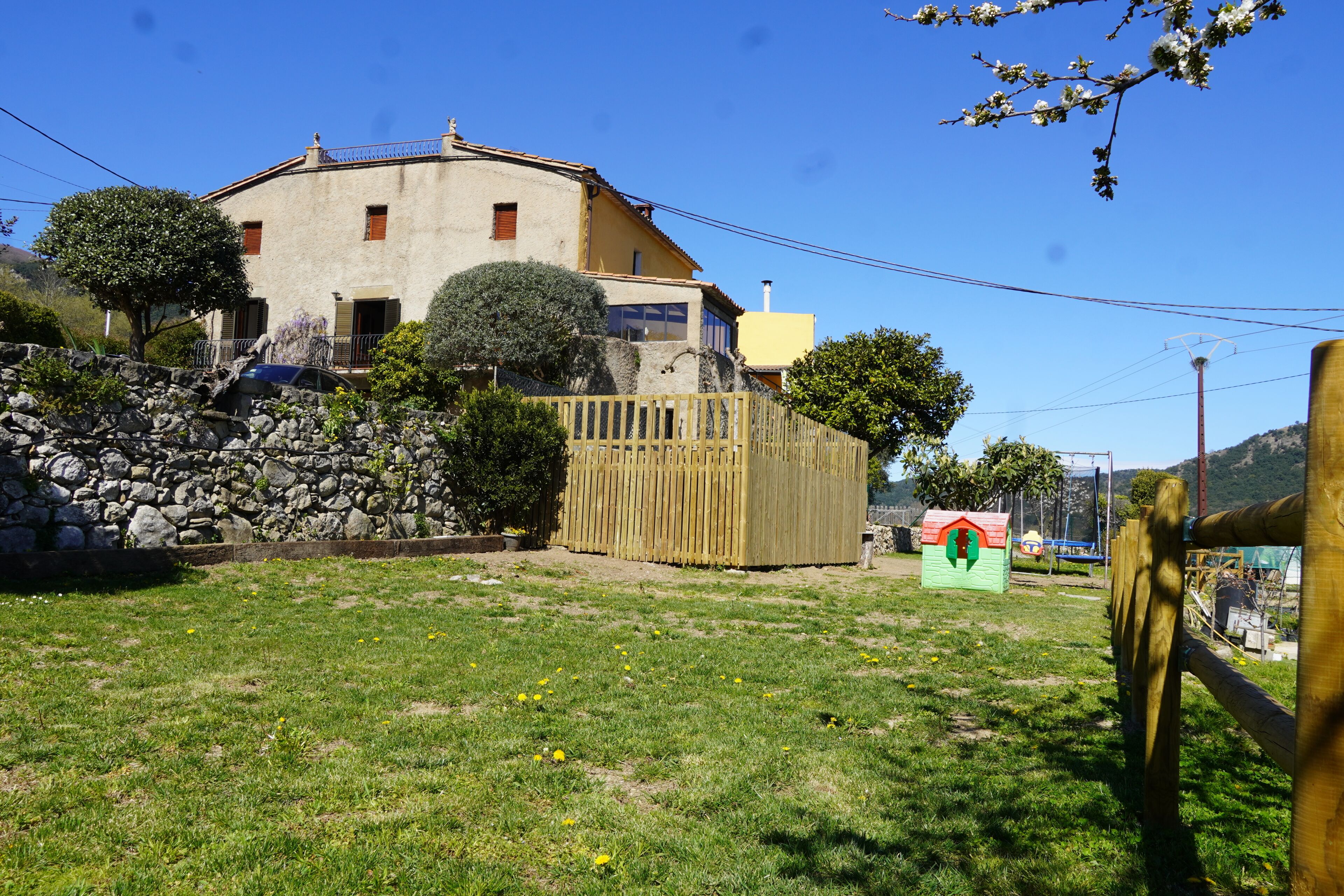 Une maison avec un mur en pierre, une clôture en bois et une petite maison de jeu dans le jardin.