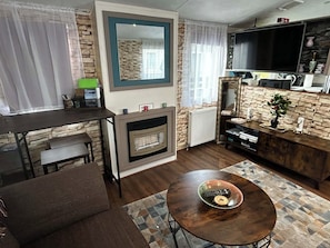 A living room with a fireplace, a television, a dining area, and a stone accent wall. - Lovely and Comfy 3-bedroom Mobile Home Retreat in Kirkcolm (Stranraer)