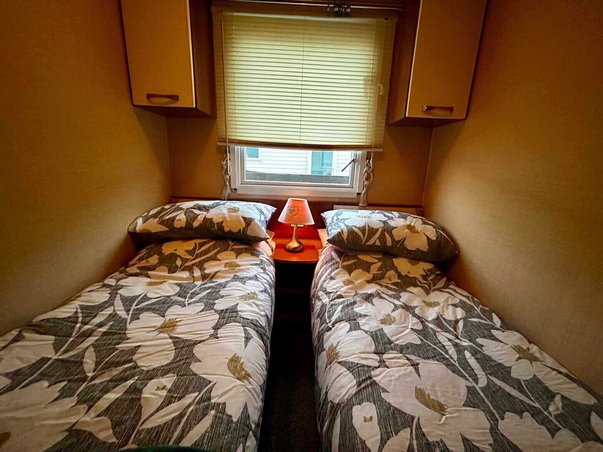 Ein kleines Zimmer mit zwei Betten, einem Fenster mit Jalousien und einem Tisch mit Lampe.