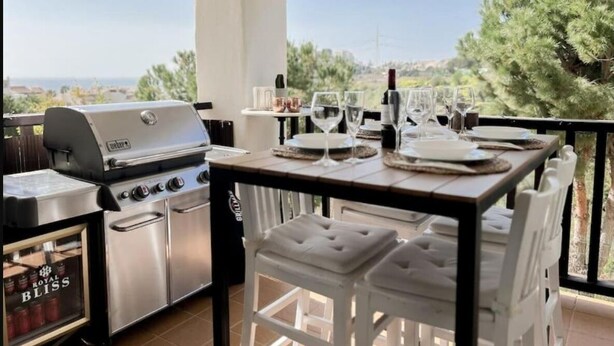 Zona d'aigües amunt per menjar a l'aire lliure amb una barbacoa, tamborets de bar blancs i una taula per a dues persones preparada amb copes de vi i plats.
