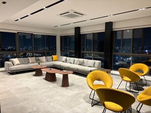 Un salon moderne avec un canapé d’angle, des tables basses rondes et des fauteuils jaunes. De grandes fenêtres offrent une vue sur la ville.