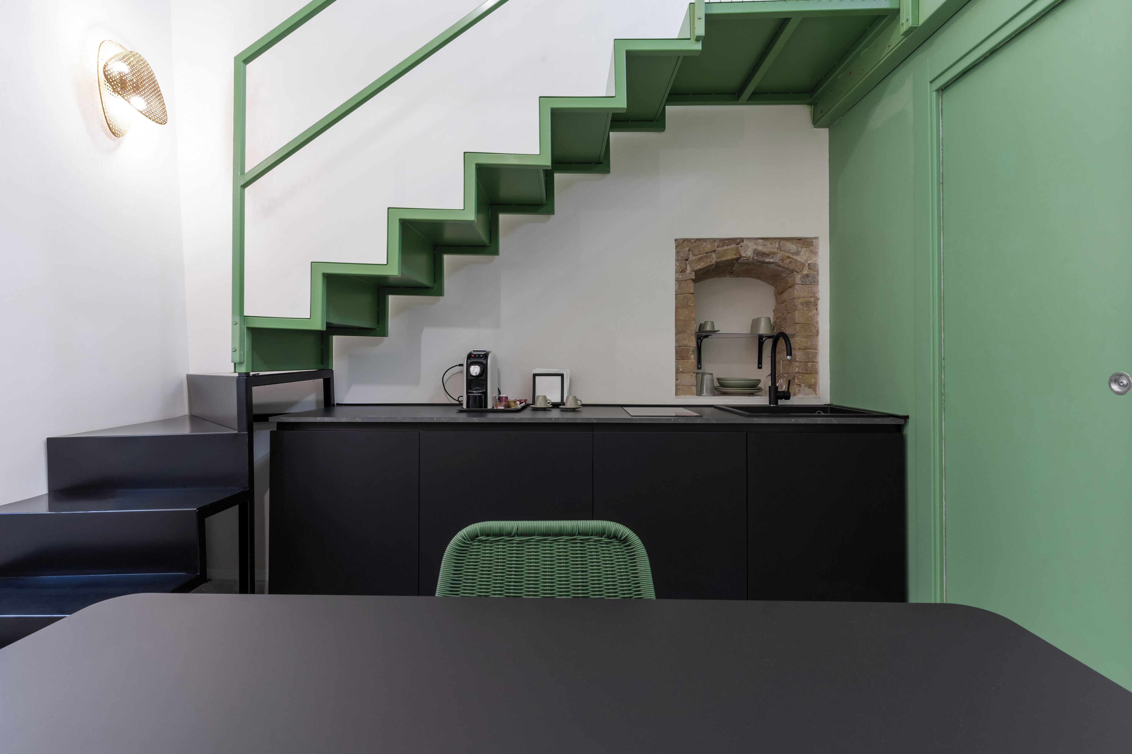 A modern kitchen with a green staircase, a black dining table, and a wicker chair.