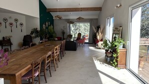 A spacious dining area with a long wooden table and chairs, a high ceiling with exposed wooden beams, and large windows providing natural light. - Group holiday in the garrigue - 12 people - total peace and quiet (Saint-Martin-de-Londres)
