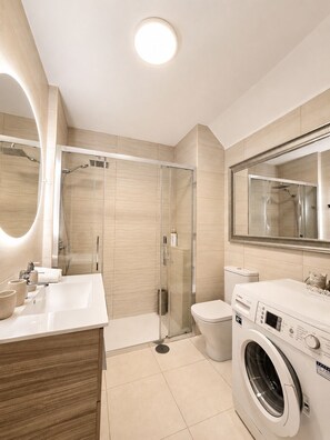 A modern bathroom with a glass-enclosed shower, a built-in washing machine, and a large mirror. - Amarilla Golf Tenerife: Apartment with Ocean View, Pool, and Terrace (Amarilla Golf)