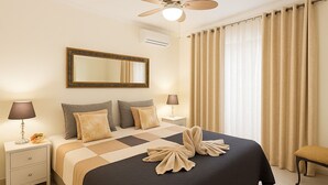 Quarto de hotel com uma cama, duas mesas de cabeceira, um ventilador de teto, um espelho grande e cortinas bege.