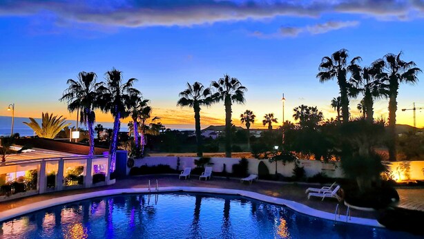 Piscina com palmeiras, pôr do sol e uma grua de construção ao longe.