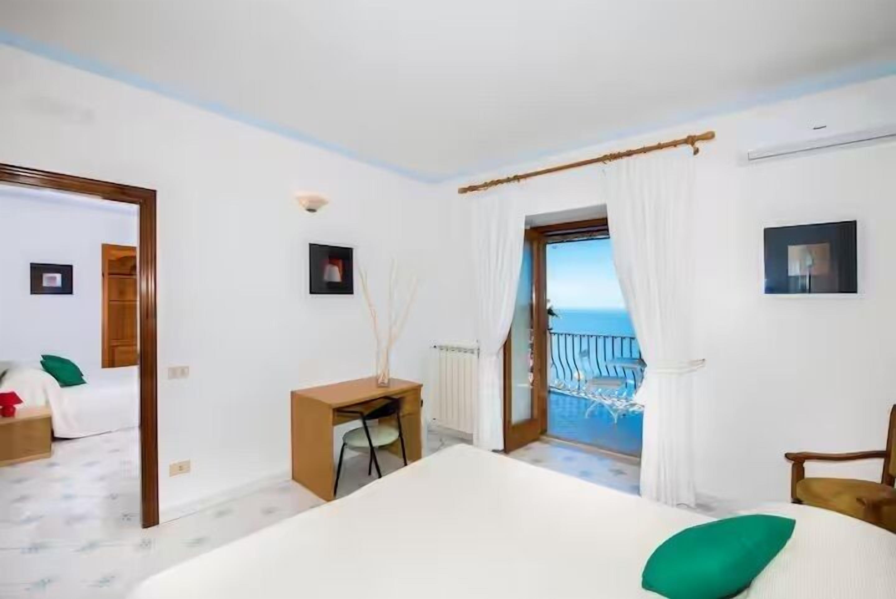 A bedroom with a bed, a desk, a chair, a radiator, a picture frame, and a view of the sea through a window.