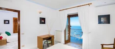 A bedroom with a bed, a desk, a chair, a radiator, a picture frame, and a view of the sea through a window.