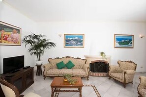 A living room with a sofa, armchairs, a coffee table, a television, a fireplace, and framed pictures on the wall. - De Vivo Realty - Villa Savina Positano (Positano)