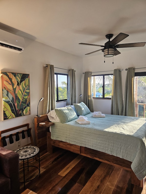 Un dormitorio con cama, ventilador de techo, escritorio y silla, ventana con cortinas y un cuadro en la pared.