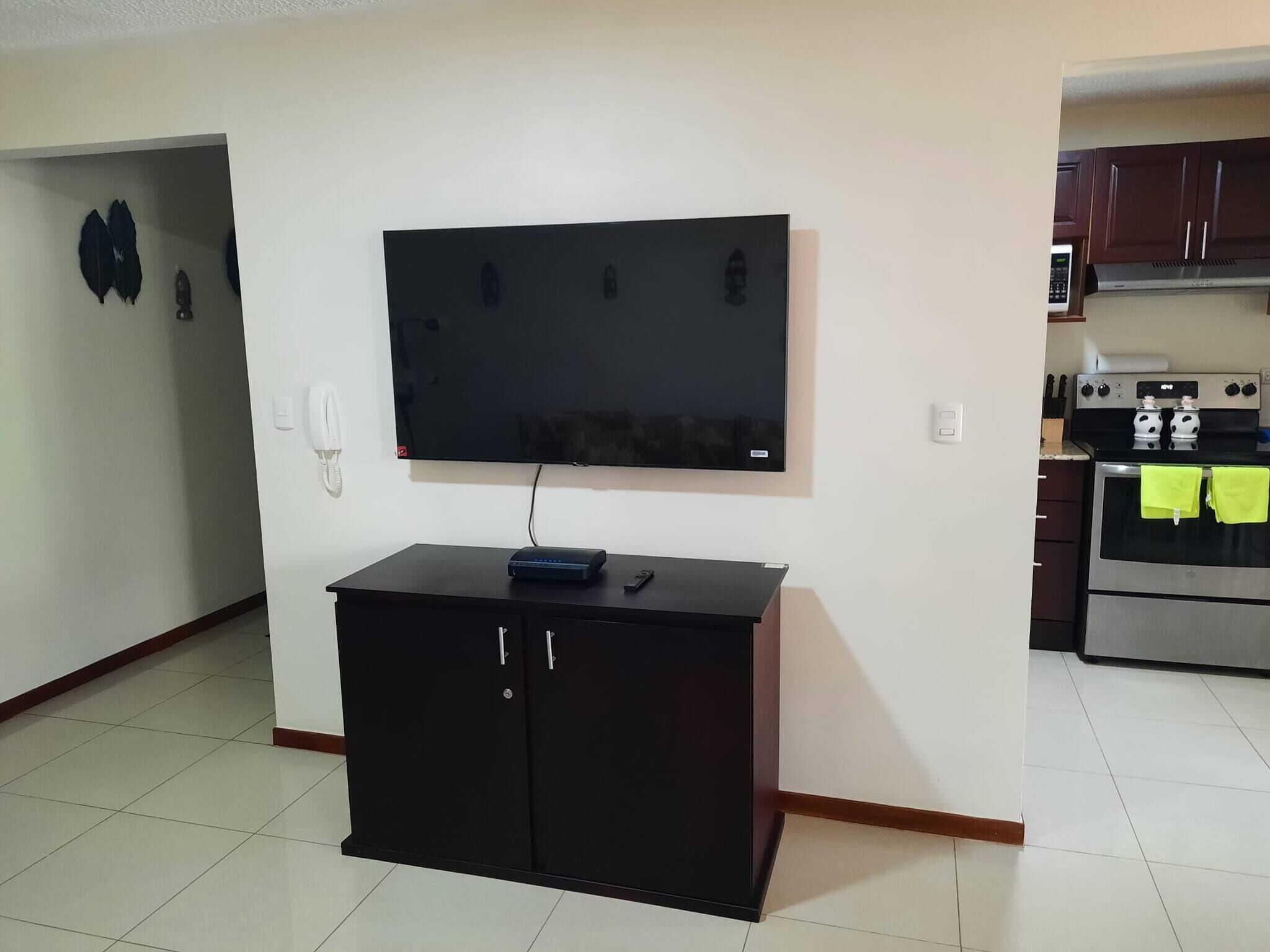 A living room with a flat-screen TV mounted on the wall, a black cabinet below, and a kitchen area with a stove and oven.