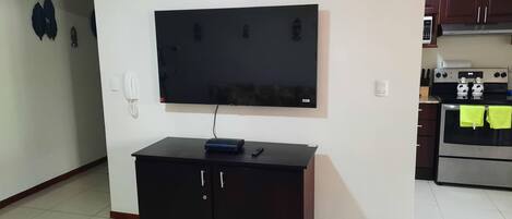 A living room with a flat-screen TV mounted on the wall, a black cabinet below, and a kitchen area with a stove and oven.