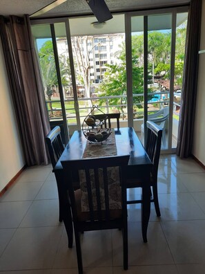 A dining area with a table and chairs, a decorative bowl, and a view of buildings and greenery through large windows. - Family-friendly Condo With Pool View, Walk to Jaco Beach (Jacó)