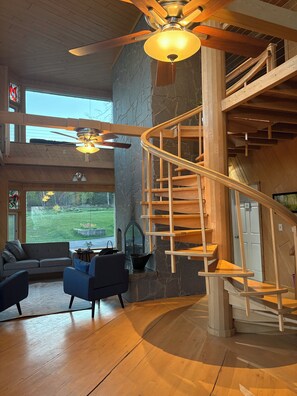 A spacious living area with a wooden staircase, a ceiling fan, and a large window offering a view of the outdoors. - Stunning Lakehouse in Castle Danger (Two Harbors)