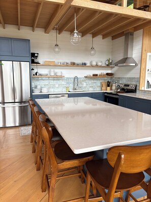 A modern kitchen with a large island, wooden chairs, stainless steel appliances, and a tiled backsplash.