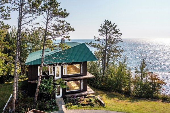 A two-story house with a green roof, large windows, and a view of the ocean.