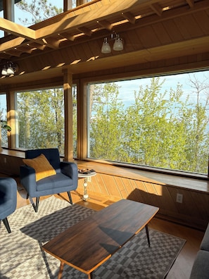 A modern interior with large windows, a wooden ceiling, a blue armchair, a wooden coffee table, and a small table with a lamp.