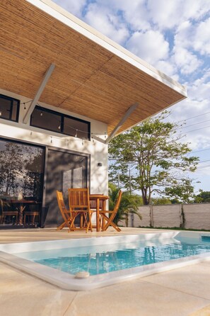 A modern outdoor pool area with a wooden deck, a covered seating area, and a large glass door leading to a building. - Private Villa with Pool & Nature (Parrita)