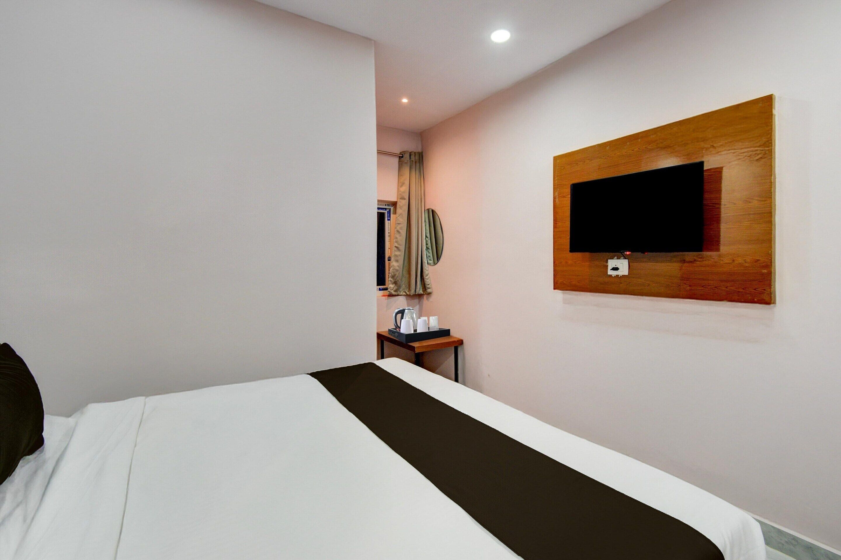 A hotel room with a bed, a TV mounted on the wall, a small table with a kettle, and a mirror.