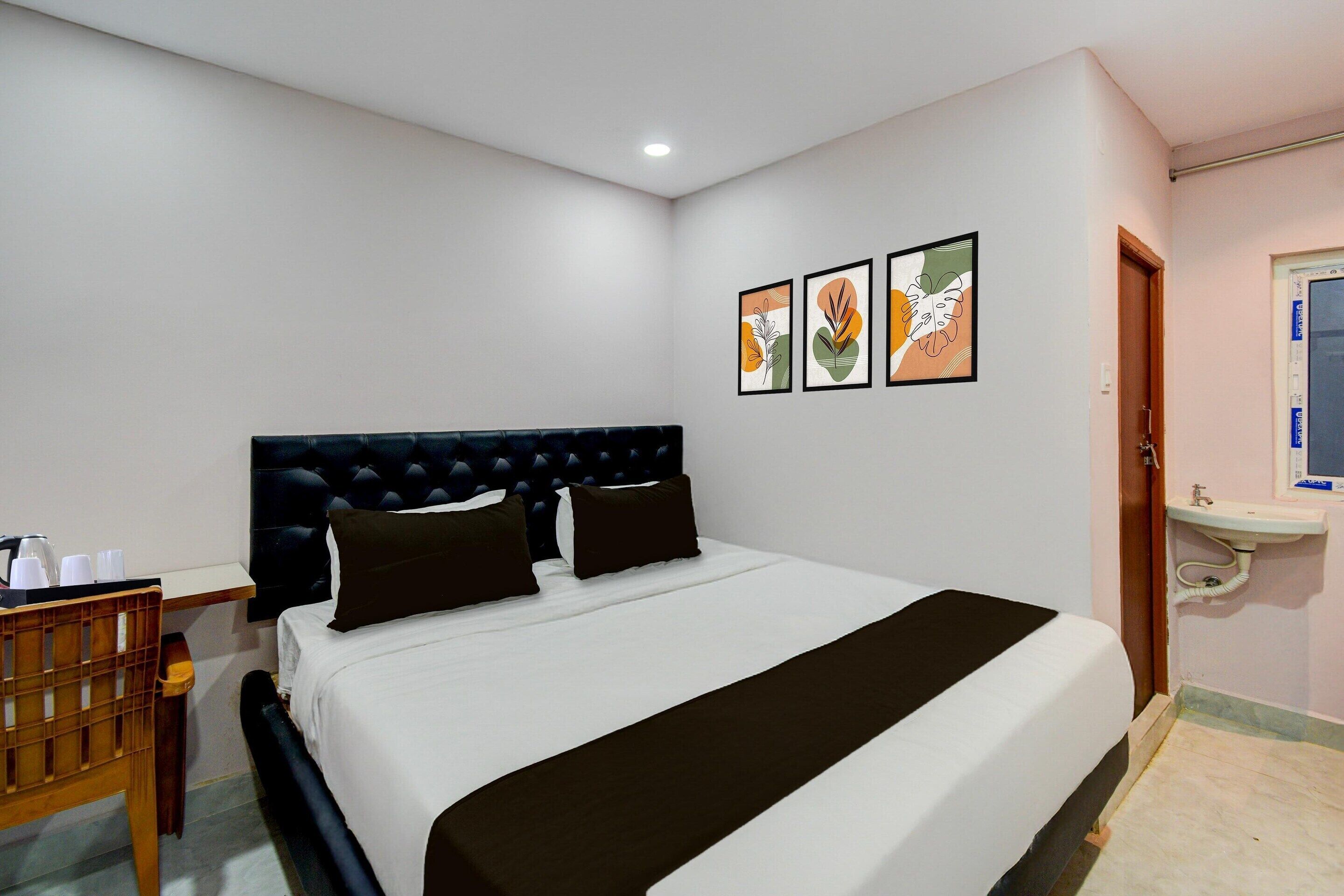 A hotel room with a bed, a wooden chair, a bedside table, a wall-mounted sink, and framed artwork.