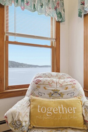 A room with a view of a lake through a window, a floral-patterned chair, and a cushion with the text 'together our favorite place to be'.
