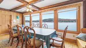 Un espace repas comprenant une table, des chaises, un ventilateur de plafond et de grandes fenêtres offrant une vue sur un plan d’eau et des collines.