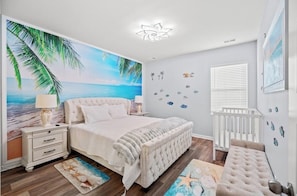 Un dormitorio con un mural con temática playera, una cuna blanca y una mesita de noche con una lámpara.