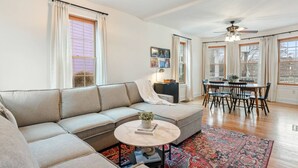 A living room with a sofa, a dining table with chairs, a ceiling fan, and large windows with curtains. - Riverfront Escape Near Boston & Transit w/ Parking (Medford)