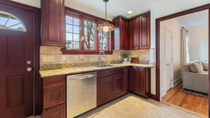 A kitchen with dark wood cabinets, a granite countertop, a stainless steel dishwasher, and a window with a view of trees. - Riverfront Escape Near Boston & Transit w/ Parking (Medford)