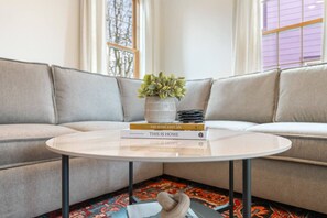 Een woonkamer met een bank, een rond bijzettafeltje met boeken en een plant, en een vloerkleed met een patroon.