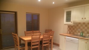 A dining area with a wooden table and chairs, a kitchen with white cabinets and a dishwasher, and a window with blinds. - STRUTHAN BEAG DINGLE (Dingle)