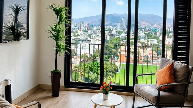 A spacious living room with a large window, a sofa, a chair, a small table, and a potted plant. -  Miraflores 1BR Apartment | Rooftop Pool, WiFi, Co-Work & City Views (Miraflores)