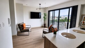 A modern living space with a flat-screen TV, a sofa, a dining area with a table, and a balcony with a city view. -  Miraflores 1BR Apartment | Rooftop Pool, WiFi, Co-Work & City Views (Miraflores)
